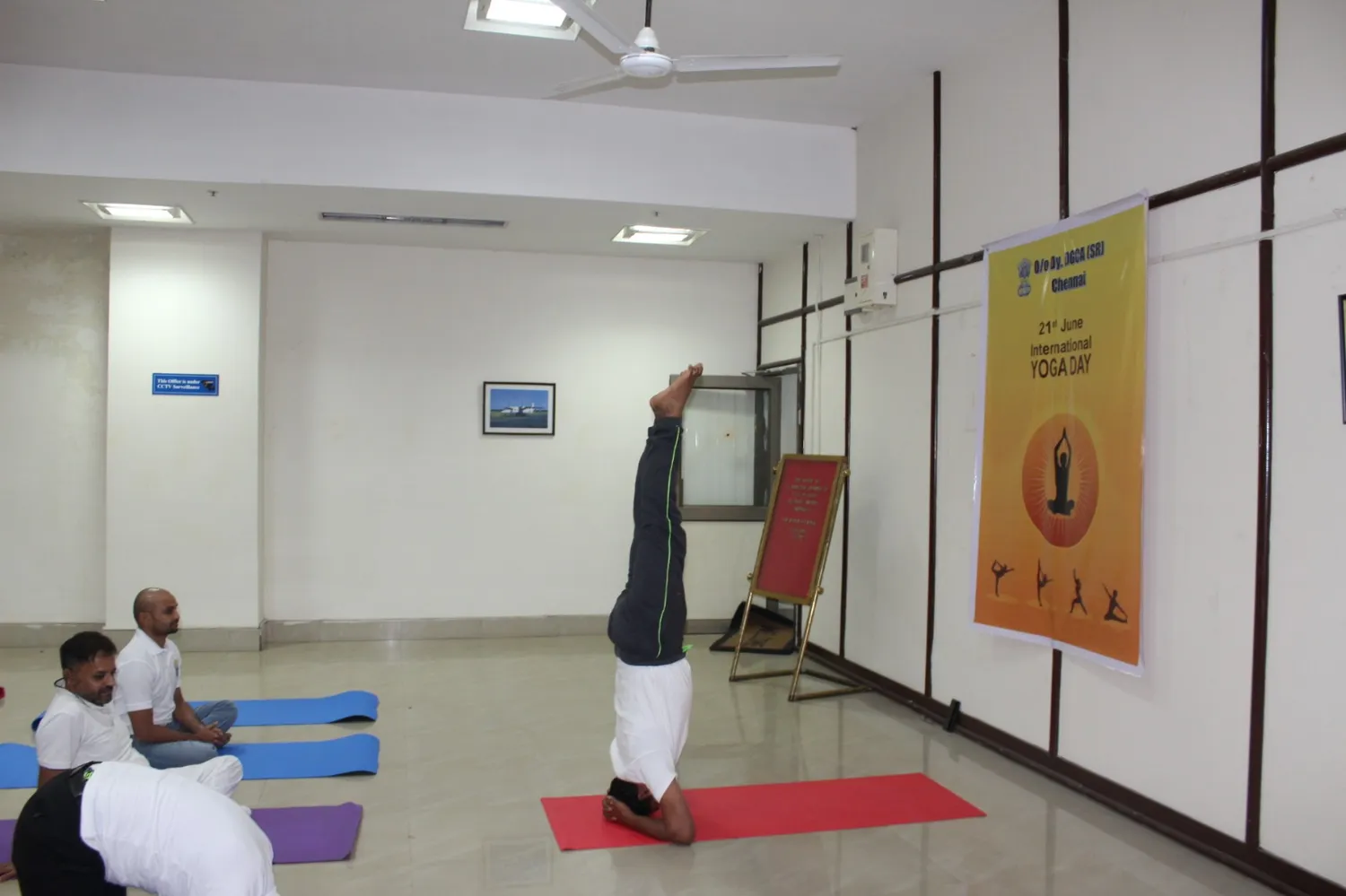 A practitioner holding a supported headstand beside an International Yoga Day banner